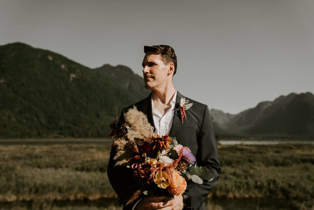 groom holding wedding bouquet with mountains background at pitt lake